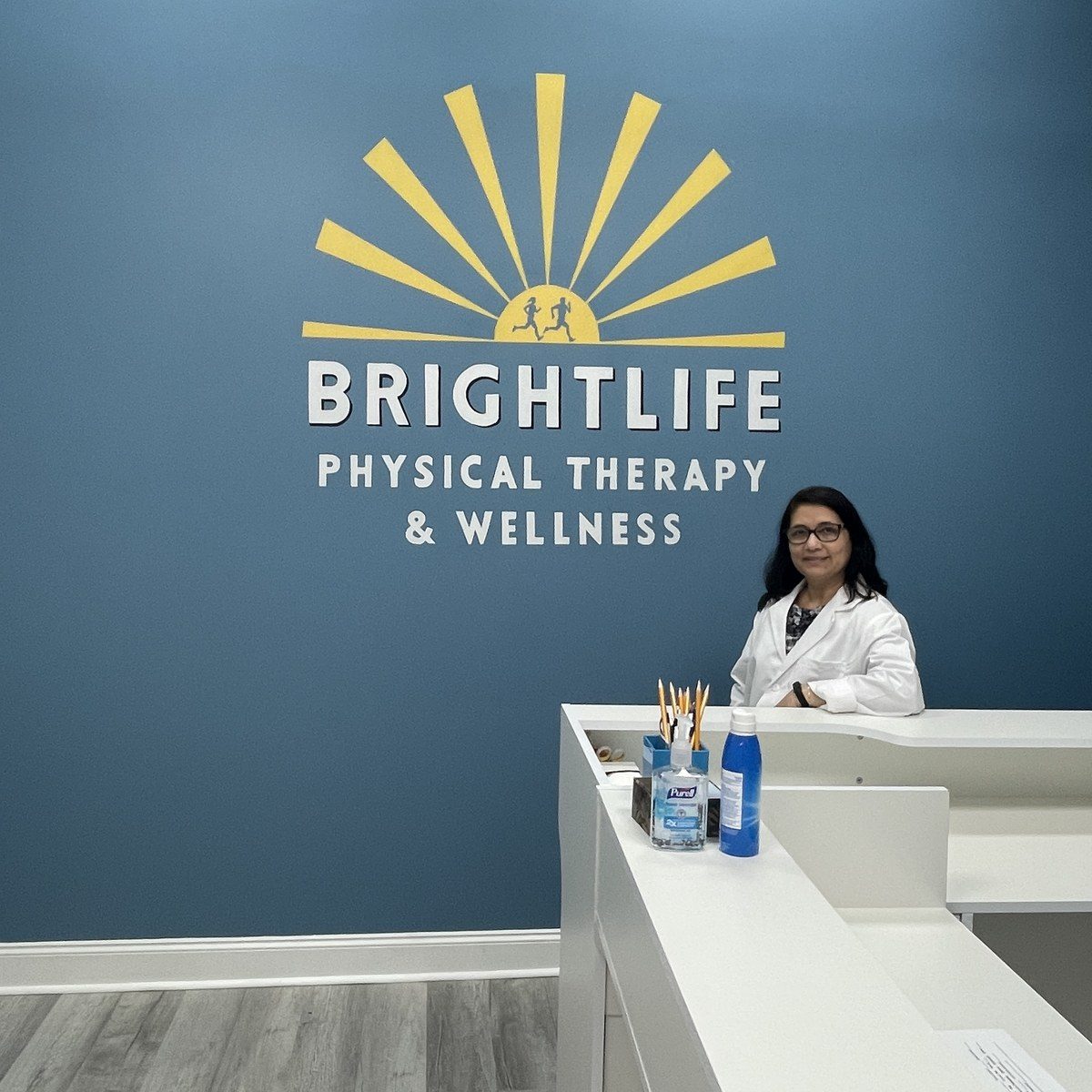 The BrightLife Physical Therapy & Wellness front desk, with the therapist welcoming patients beneath the sunrise logo mural on the clinic's blue accent wall