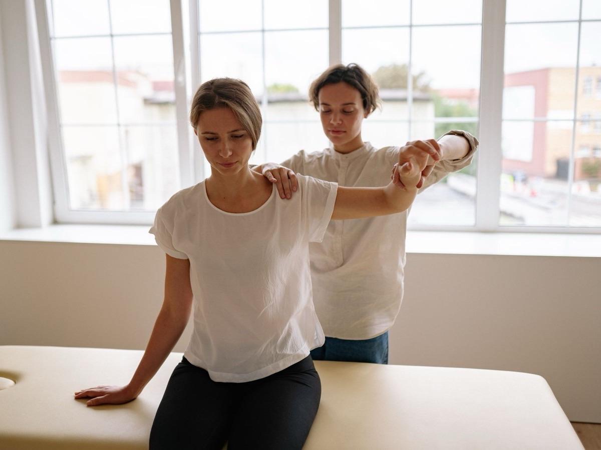 Physical therapist guiding a seated patient's arm and shoulder through hands-on treatment at a therapy table