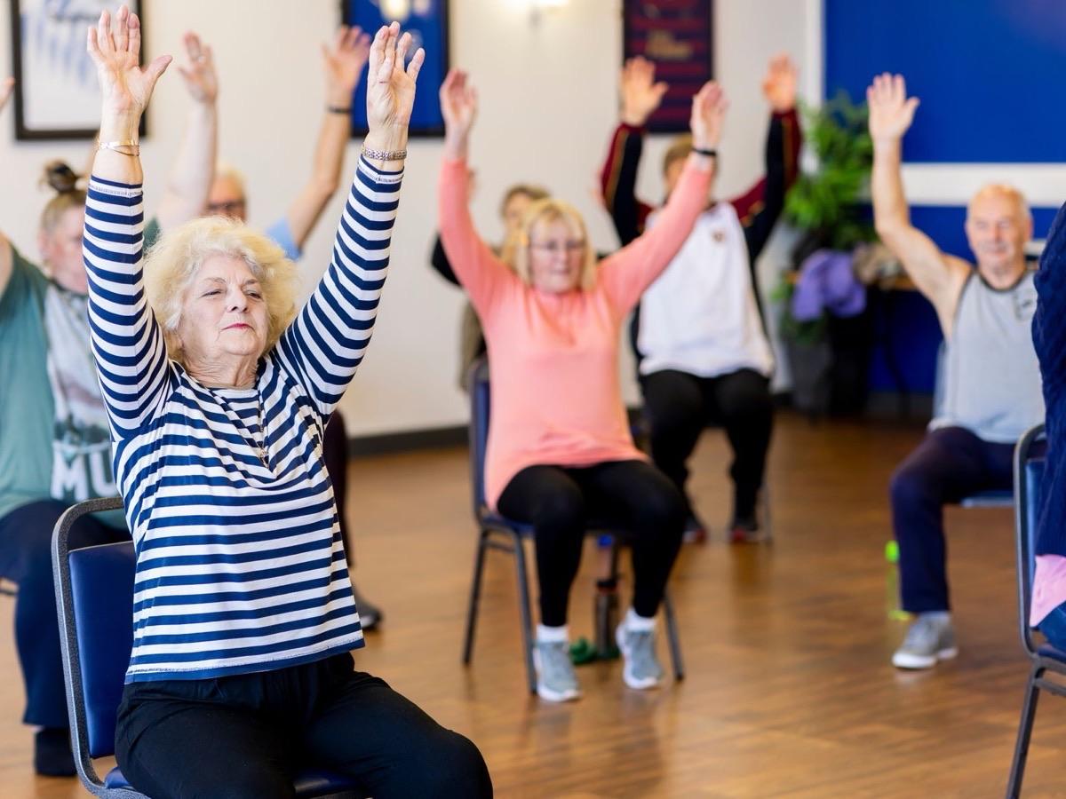 A cheerful group of seniors raising their arms overhead during a seated group exercise class
